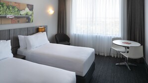 A hotel room with two beds, a small table, a chair, a lamp, and a large window with curtains. - Mercure Sydney Bankstown (Bankstown)