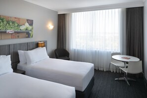 A hotel room with two beds, a small table, a chair, a lamp, and a large window with curtains. - Mercure Sydney Bankstown (Bankstown)