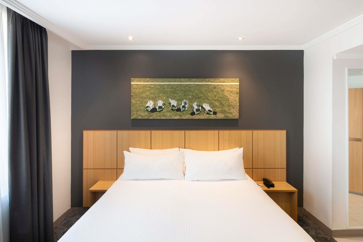 A modern hotel room with a large bed, wooden headboard, and a painting of gloves on the wall.