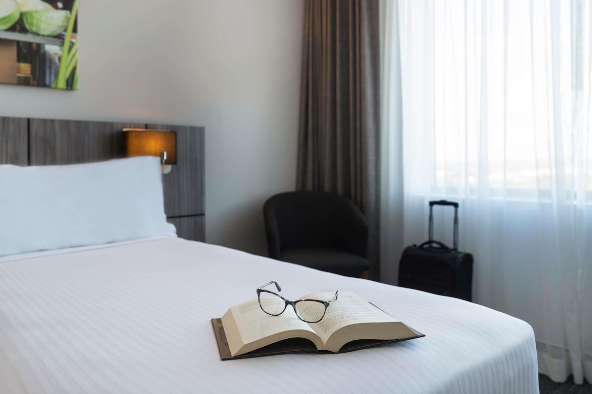 A hotel room with a bed, a nightstand with a lamp, a chair, a suitcase, and an open book with glasses on it.