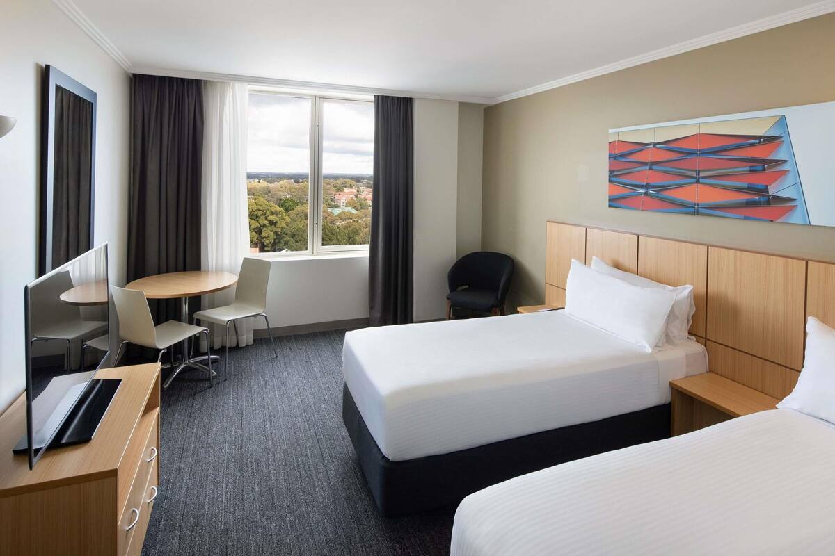 A hotel room with two beds, a desk, a chair, a TV, and a large window with a view.