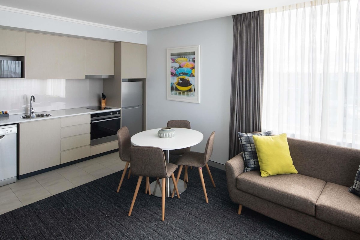 A modern hotel room with a kitchenette, a dining area with a round table and chairs, a sofa with cushions, and a large window with curtains.