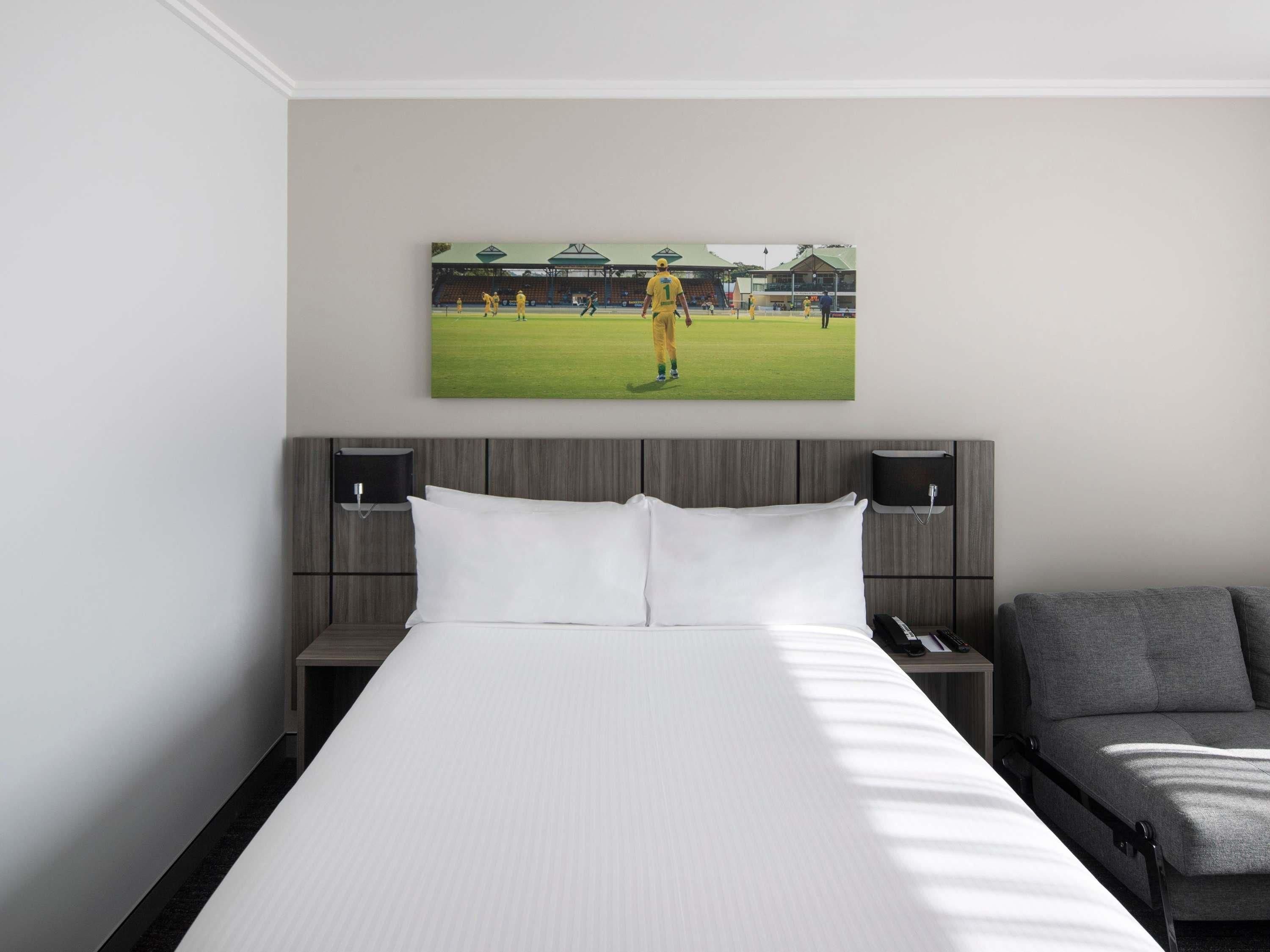 Hotelkamer met een groot bed, twee nachtkastjes, een grijze bank en een aan de muur hangend beeld van een sportveld.