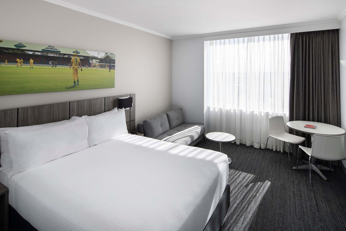 A hotel room with a large bed, a sofa, a small table, and a chair. There is a painting of a soccer field on the wall.