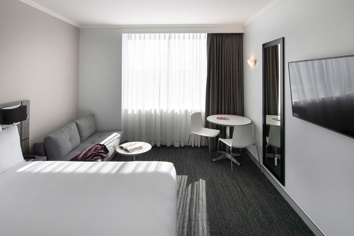 A hotel room with a bed, a sofa, a small table, a chair, a TV, and a window with curtains.