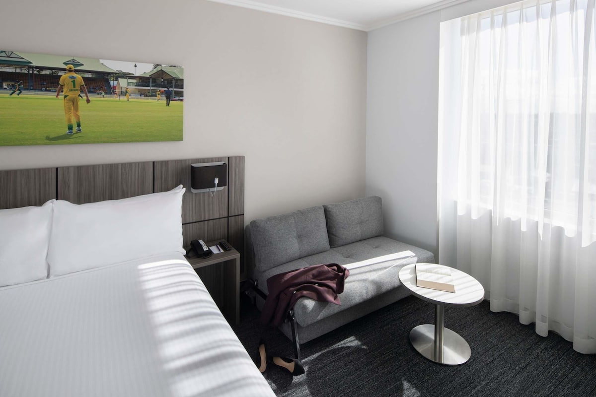 A hotel room with a bed, a grey sofa, a small round table, a TV, and a window with sheer curtains.