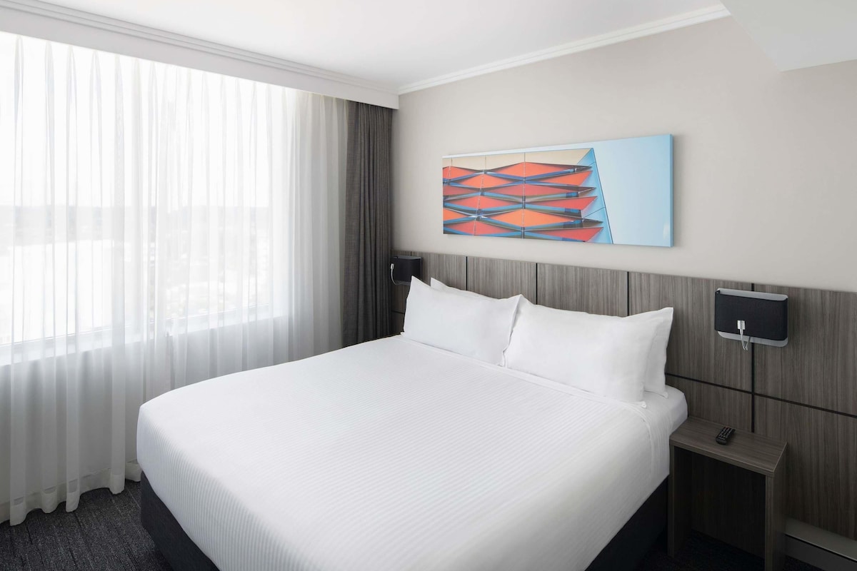 A hotel room with a large bed, a nightstand, a wall-mounted TV, and a window with sheer curtains.