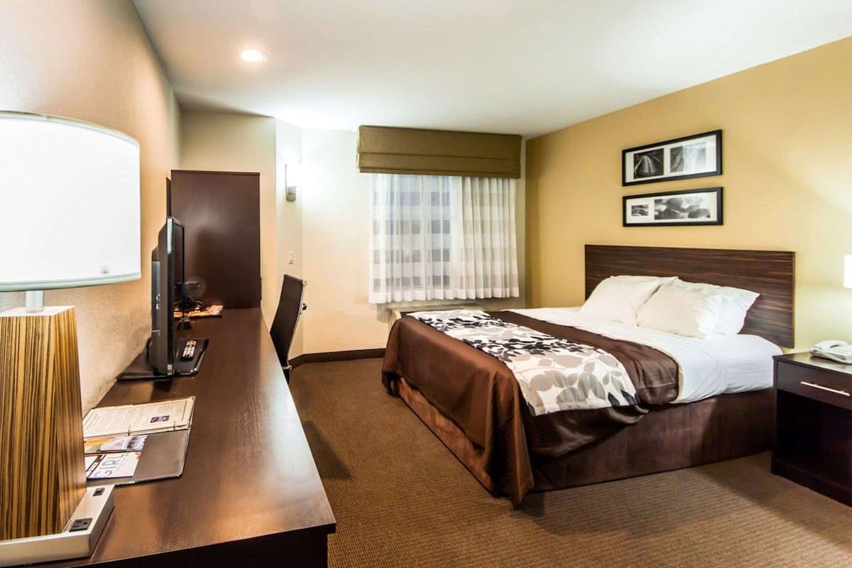 Ett hotellrum med en säng, ett skrivbord med en lampa, en tv och ett fönster med gardiner.