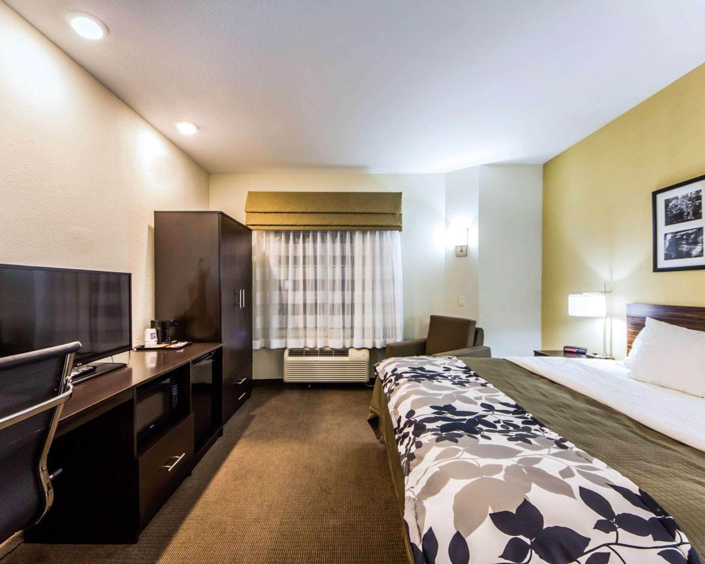 A hotel room with a bed, a desk with a television, a wardrobe, an air conditioning unit, and a window with curtains.