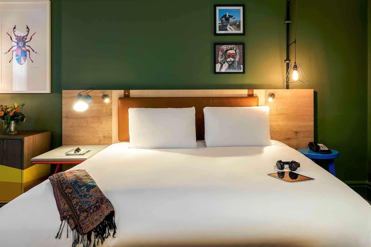 Ett hotellrum med en stor säng, en sänggavel i trä, två sängbord, ett litet bord, en lampa och konstverk på väggen.