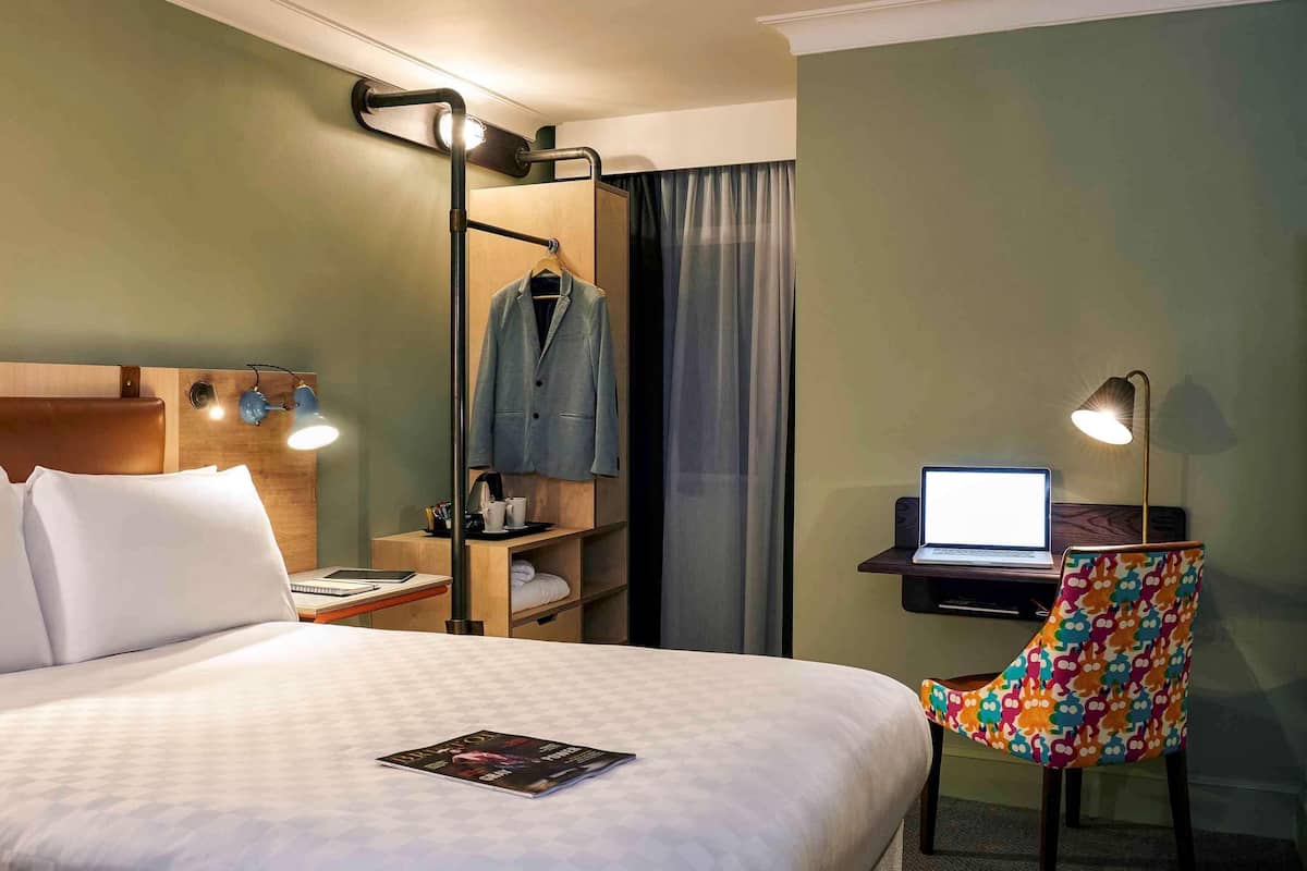 Ett hotellrum med en säng, ett skrivbord med en bärbar dator, en stol, en klädställning och en hylla med en jacka.