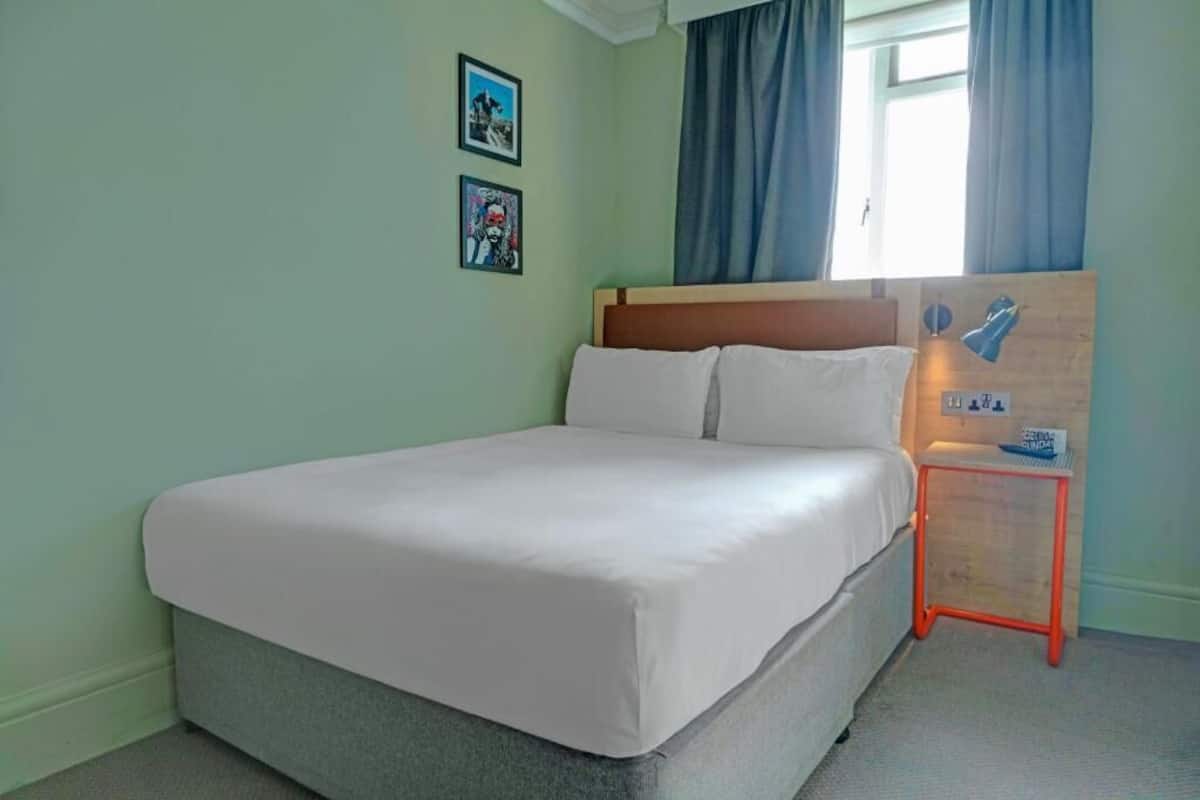 Een net opgemaakt bed in een kamer met lichtgroene wanden, twee ingelijste foto’s aan de muur, een raam met blauwe gordijnen en een klein oranje tafeltje met een lamp.