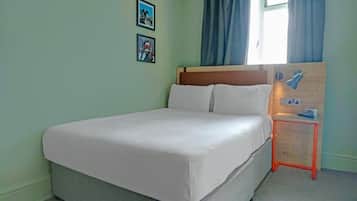 Una cama bien hecha en una habitación con paredes verde claro, dos cuadros en la pared, una ventana con cortinas azules y una mesita naranja con una lámpara.