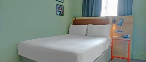 Un letto rifatto con cura in una stanza dalle pareti verde chiaro, due quadri appesi alla parete, una finestra con tende blu e un piccolo tavolino con una lampada.