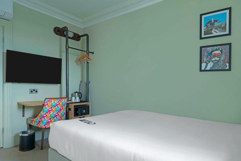 A hotel room with a bed, a desk, a chair, a coat rack, and two framed pictures on the wall.