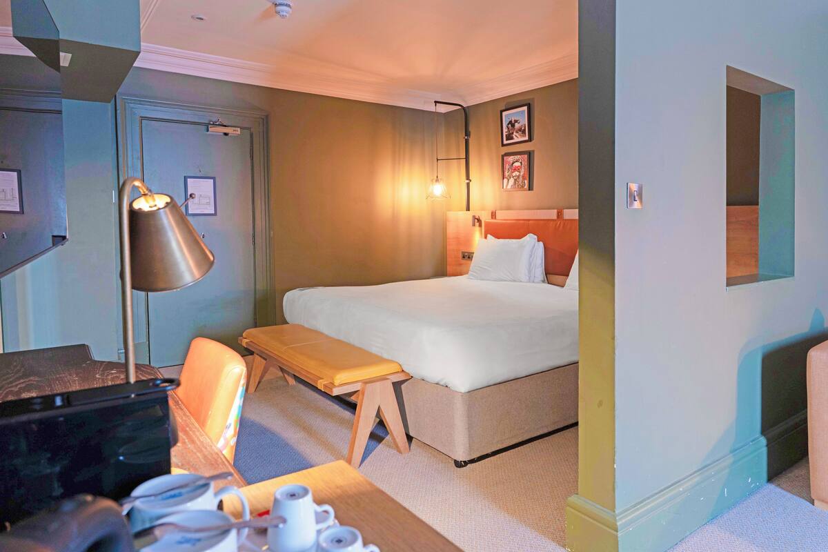 Een hotelkamer met een bed, nachtkastje, lamp en een bureau met stoel.