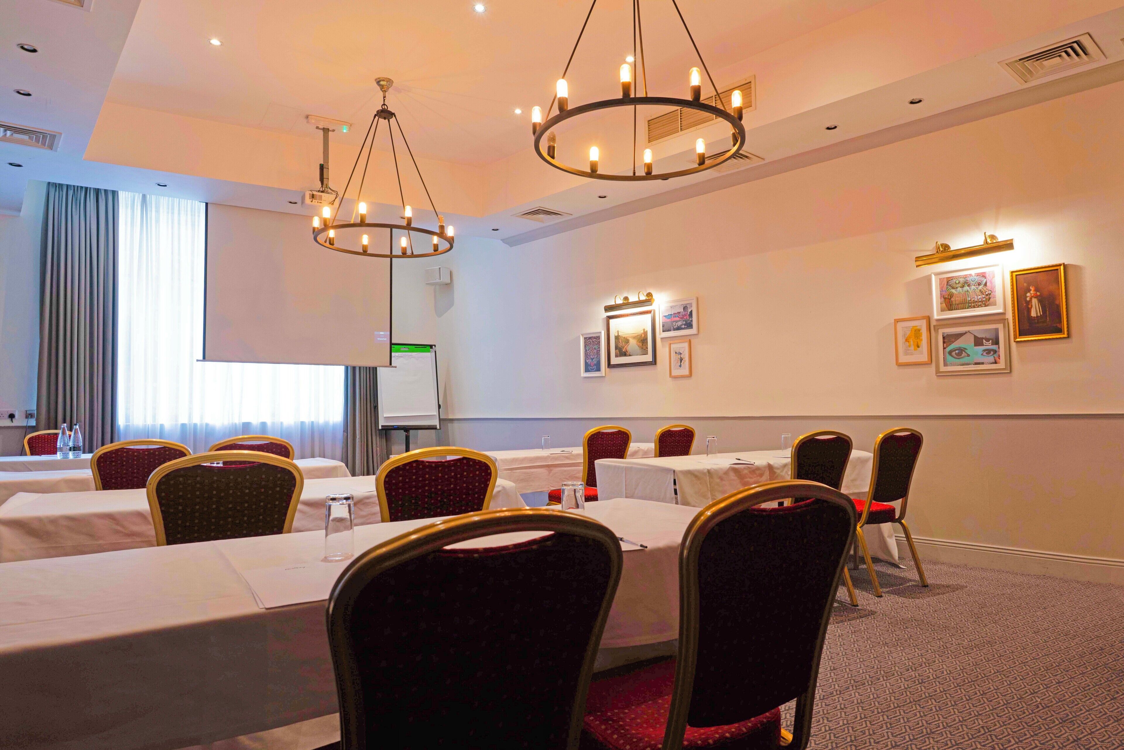 A conference room with multiple tables and chairs, a projector screen, and framed artwork on the walls.