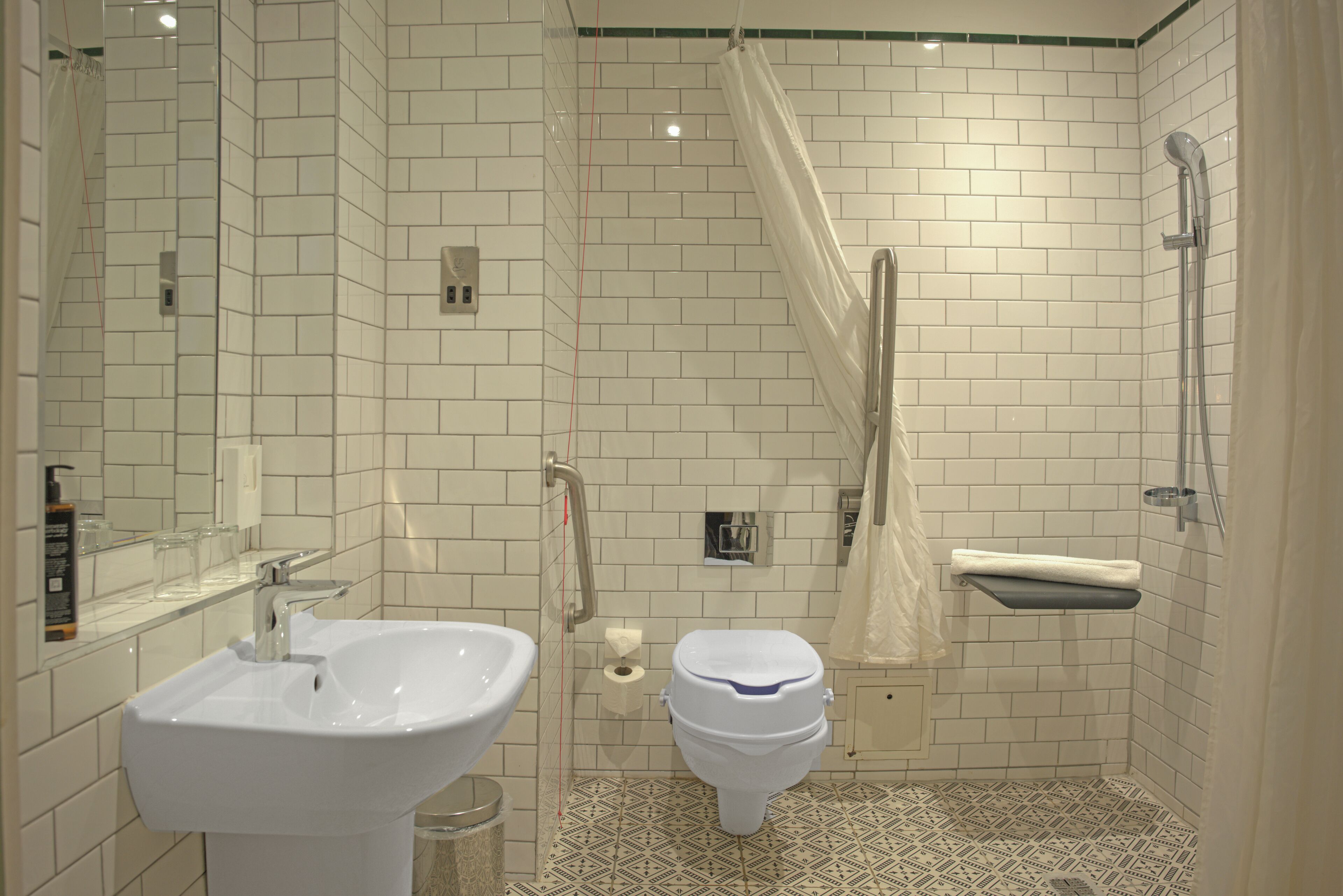 A bathroom with a white sink, toilet, and shower area, featuring white tiled walls and a patterned floor.