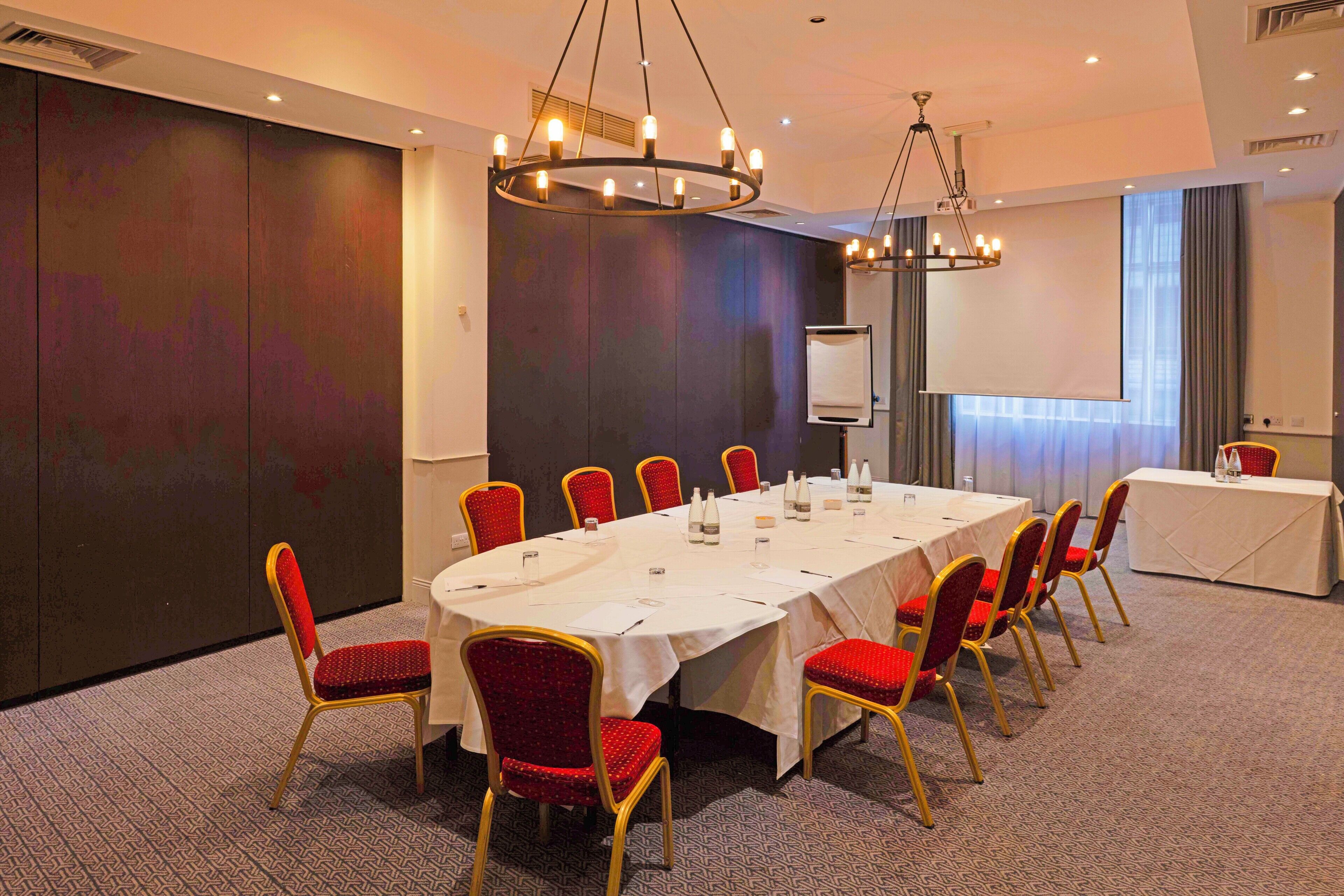 A conference room with a long table, red chairs, and a projector screen.