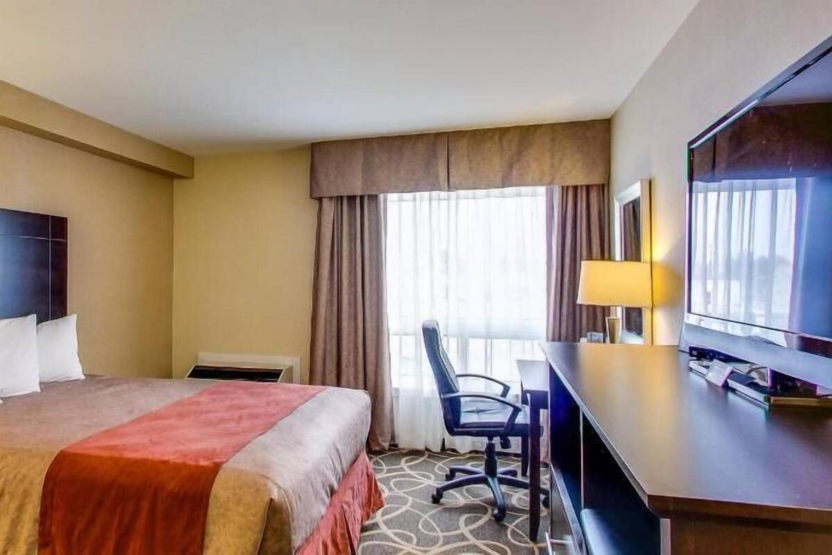 Habitación de hotel con cama, escritorio, silla, televisor y ventana grande con cortinas.