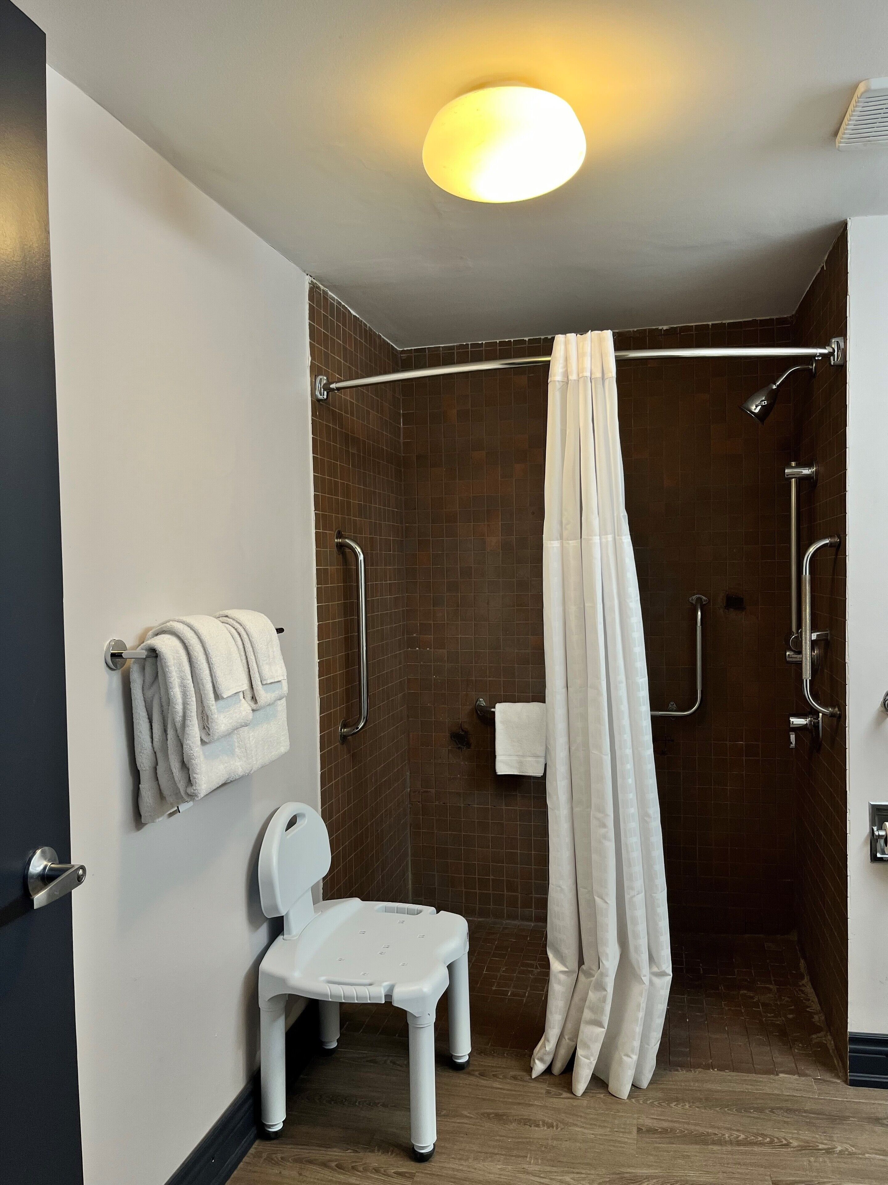A modern bathroom with a shower, grab bars, a white chair, and towels on a rack.