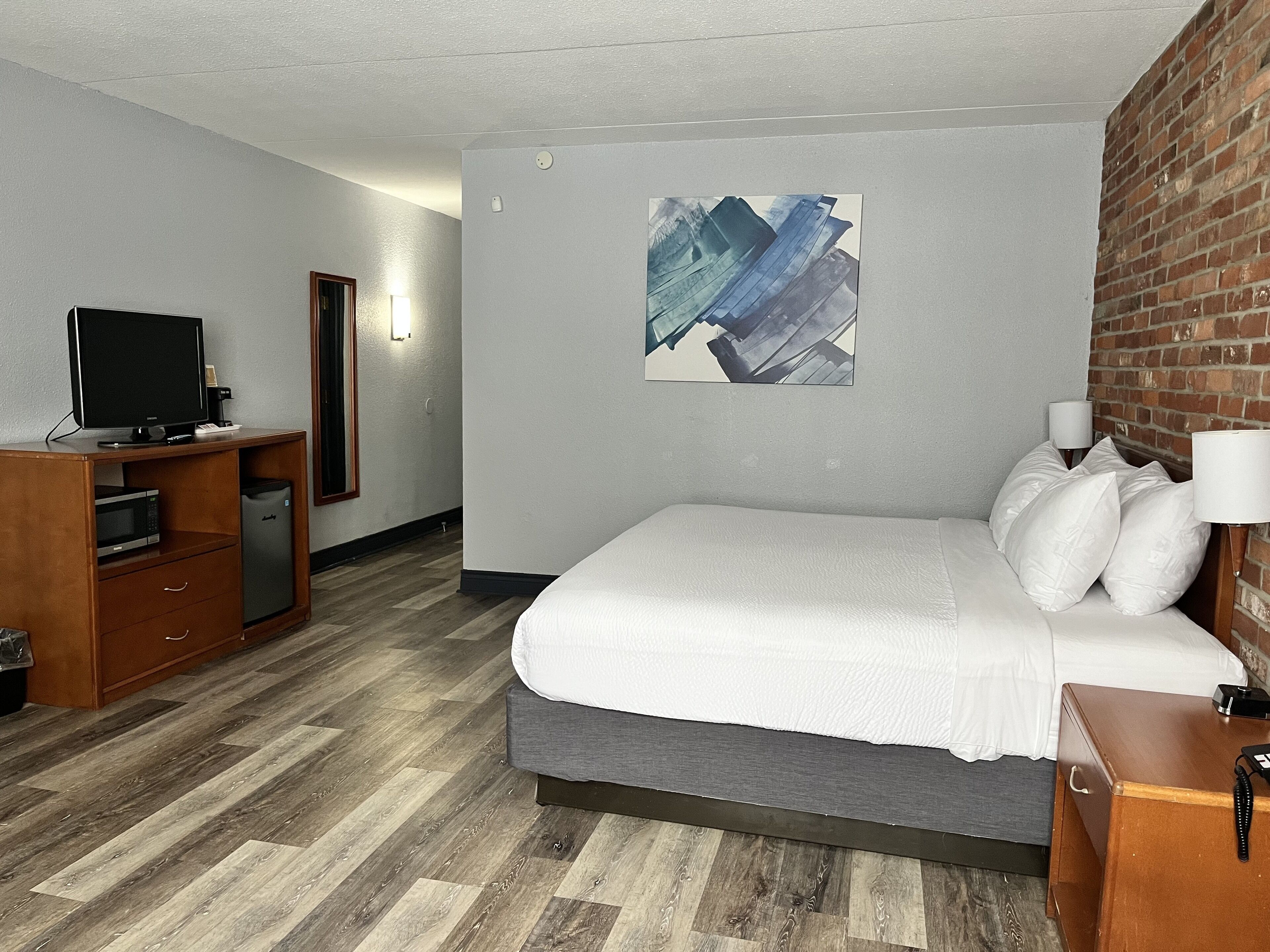 Quarto de hotel com uma cama, uma escrivaninha com um computador, uma televisão e uma parede de tijolos.