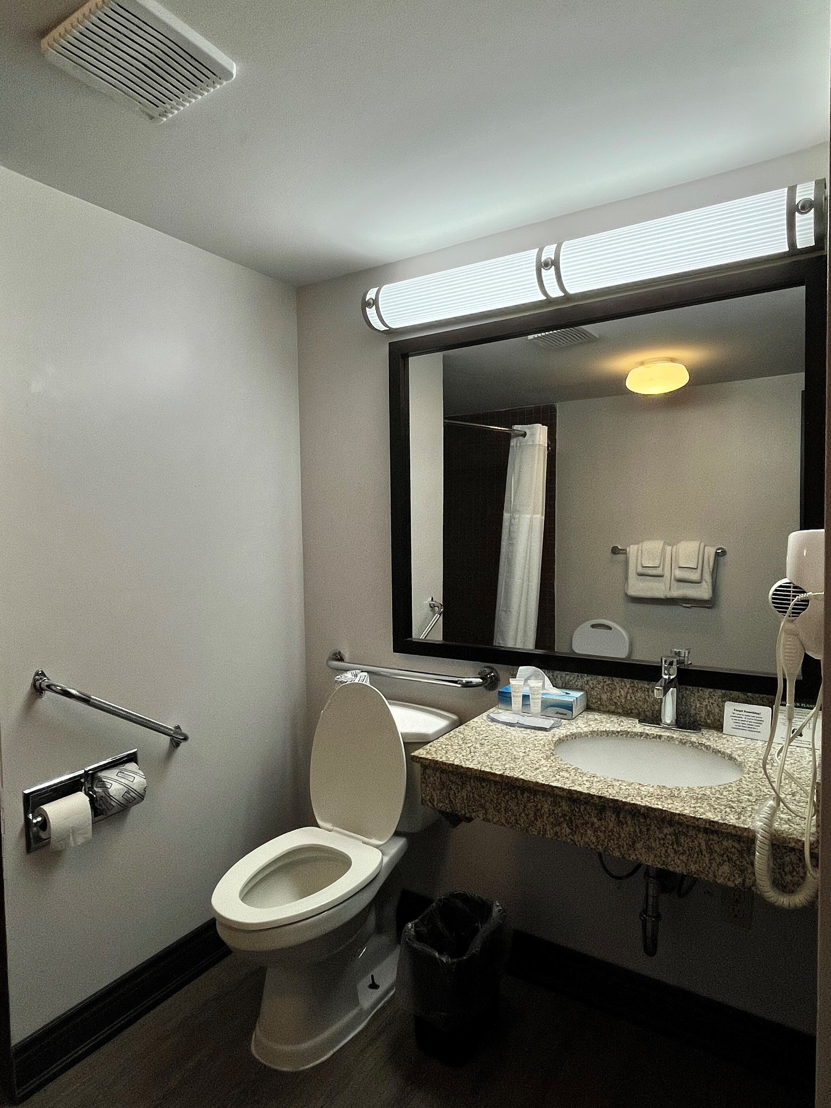 Une salle de bain d’hôtel standard, équipée de toilettes, d’un lavabo, d’un miroir, d’un porte-serviettes et d’une poubelle.
