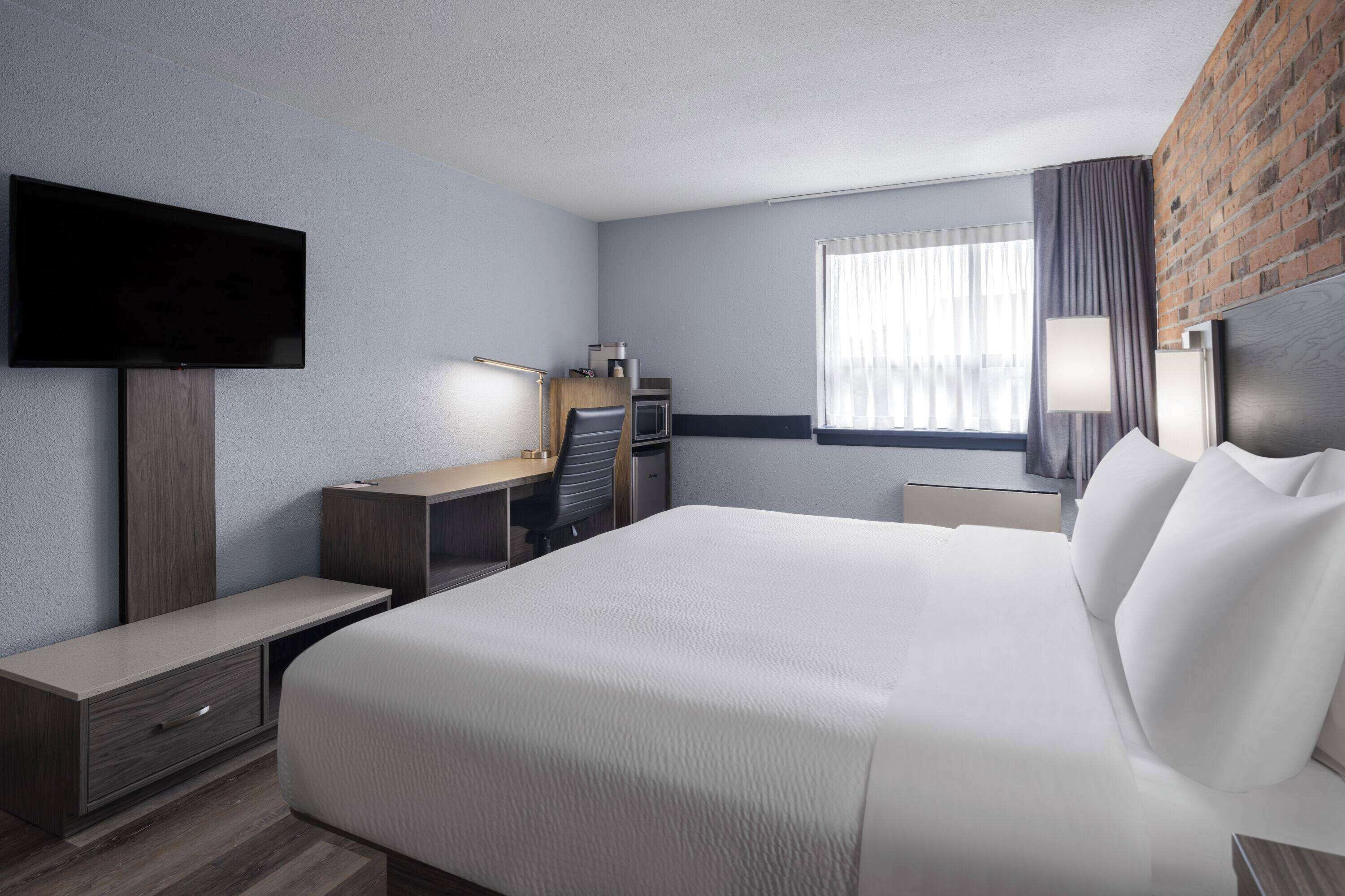 Een hotelkamer met een groot bed, een bureau met stoel, een televisie, een raam met gordijnen en een bakstenen muur.
