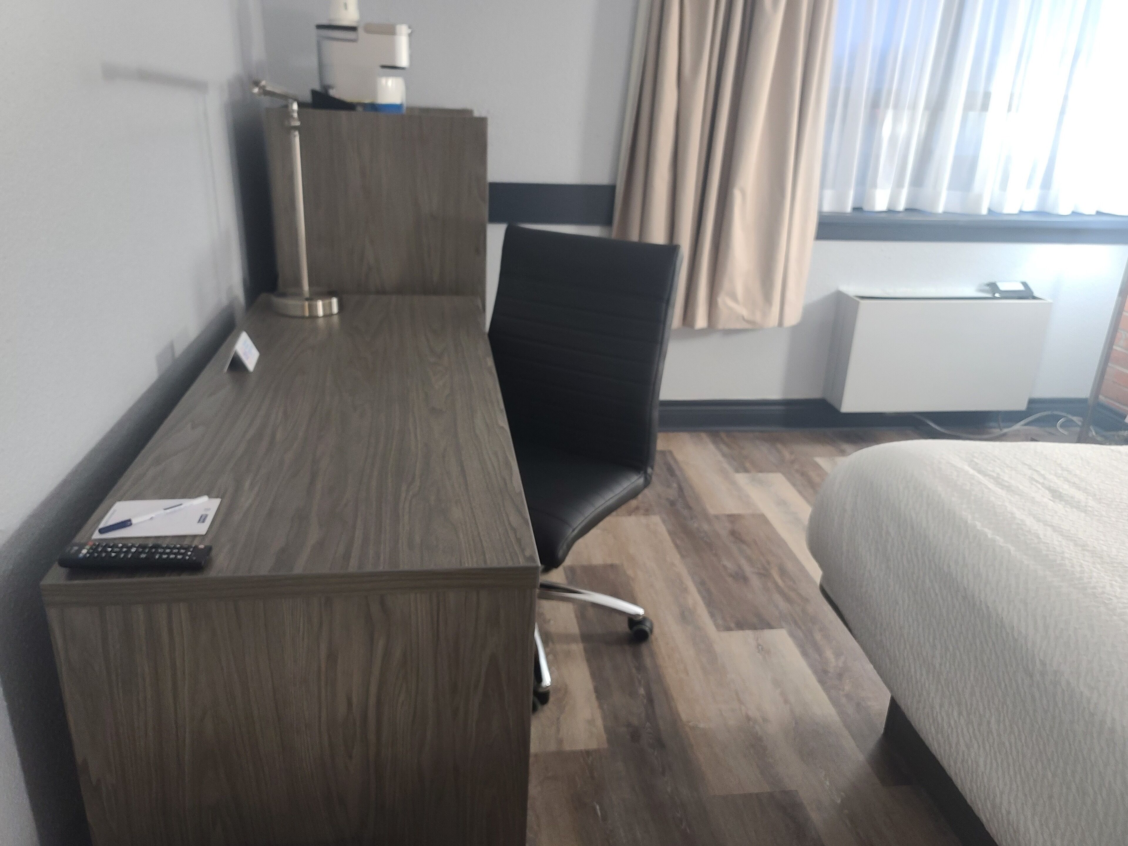 Una habitación de hotel con un escritorio de madera, una silla de oficina negra, una cama y una ventana con cortinas.