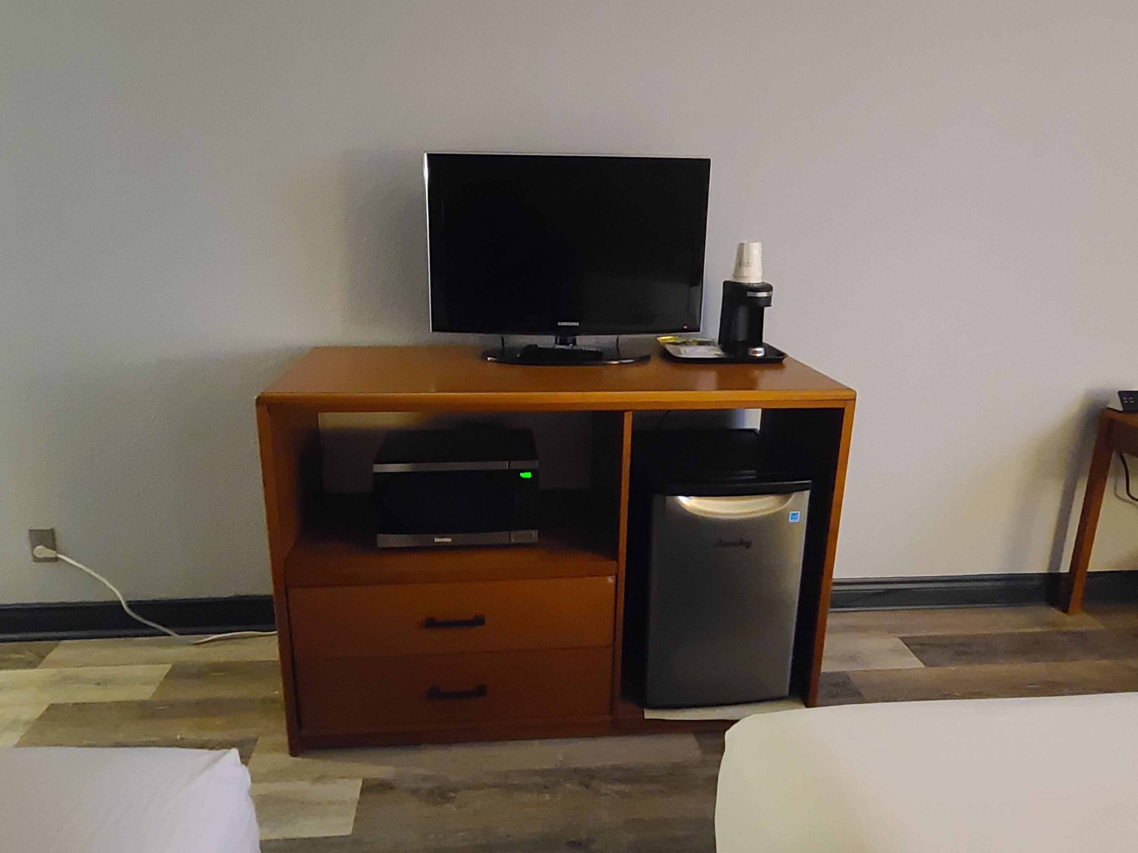 A hotel room with a wooden desk, a flat-screen TV, a coffee maker, a printer, and a refrigerator.