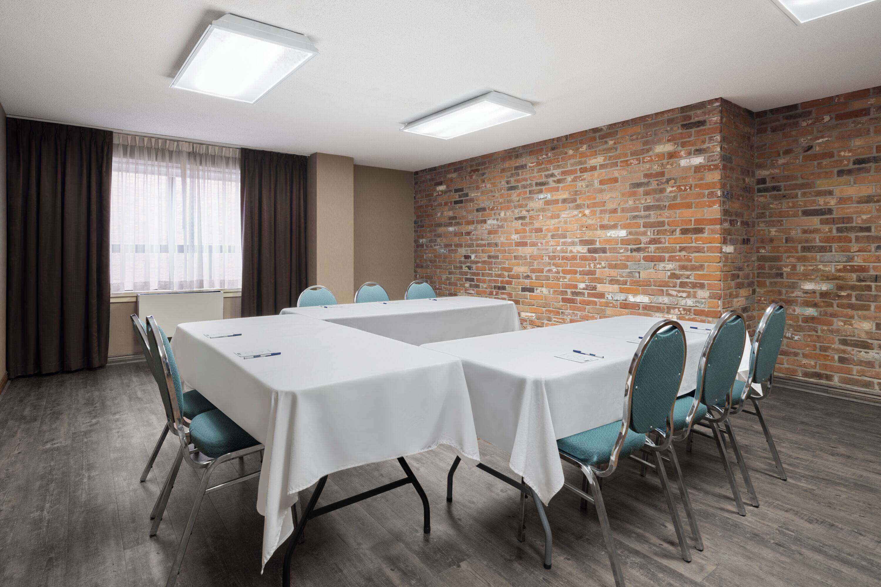 A conference room with two tables, chairs, a brick wall, and a window with curtains.