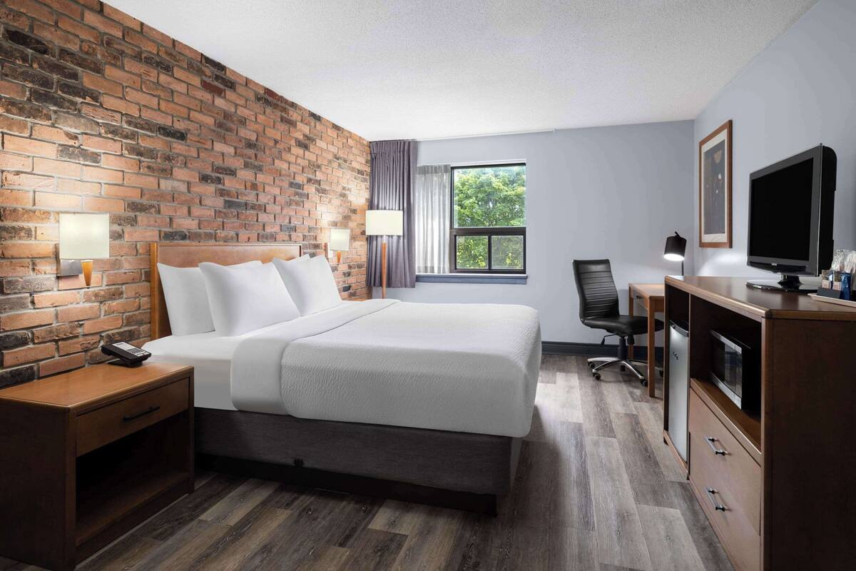 Une chambre d’hôtel avec un mur en briques, un grand lit, un bureau avec une télévision et une fenêtre donnant sur des arbres.
