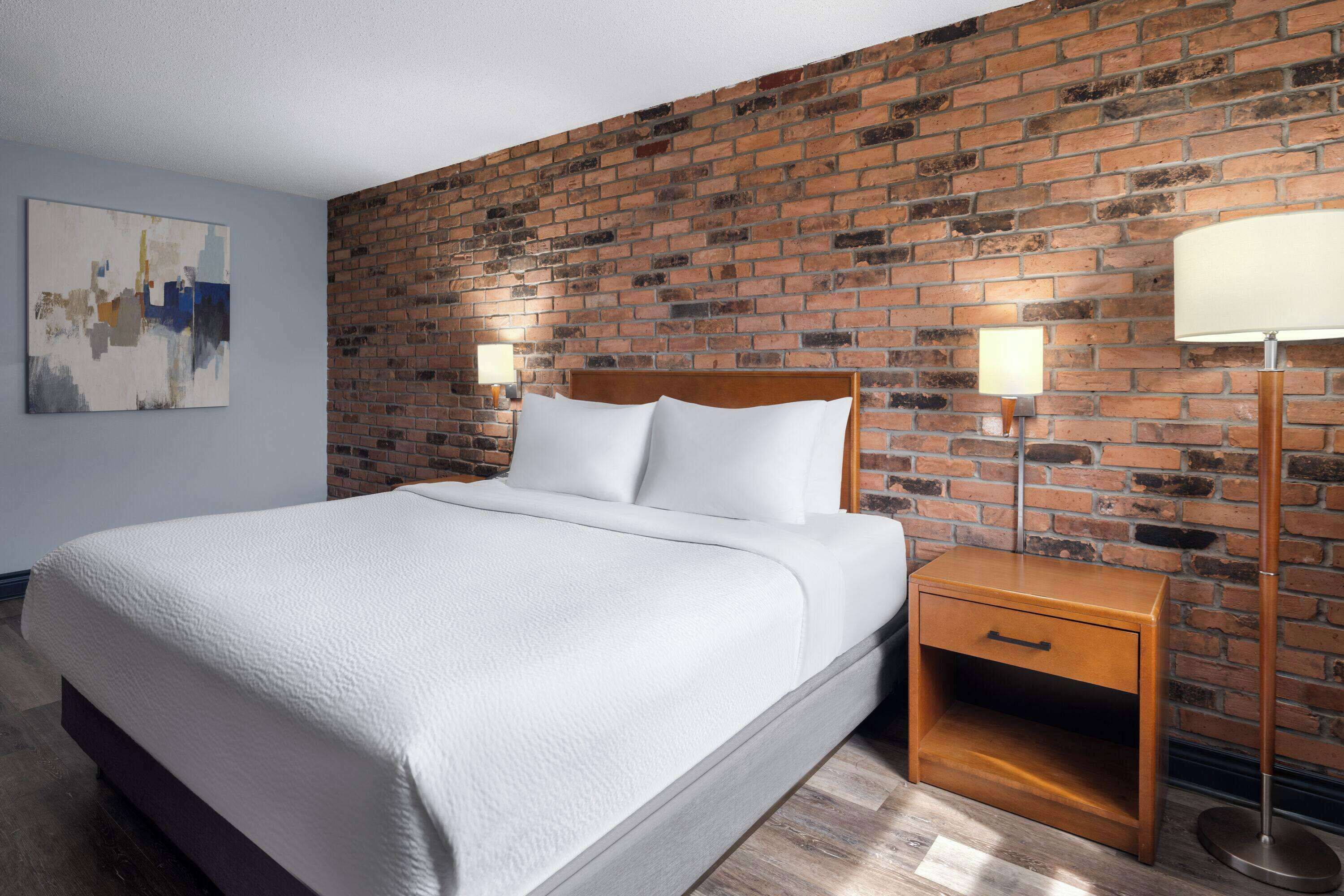 A hotel room with a brick wall, a bed with white bedding, a nightstand, and a floor lamp.