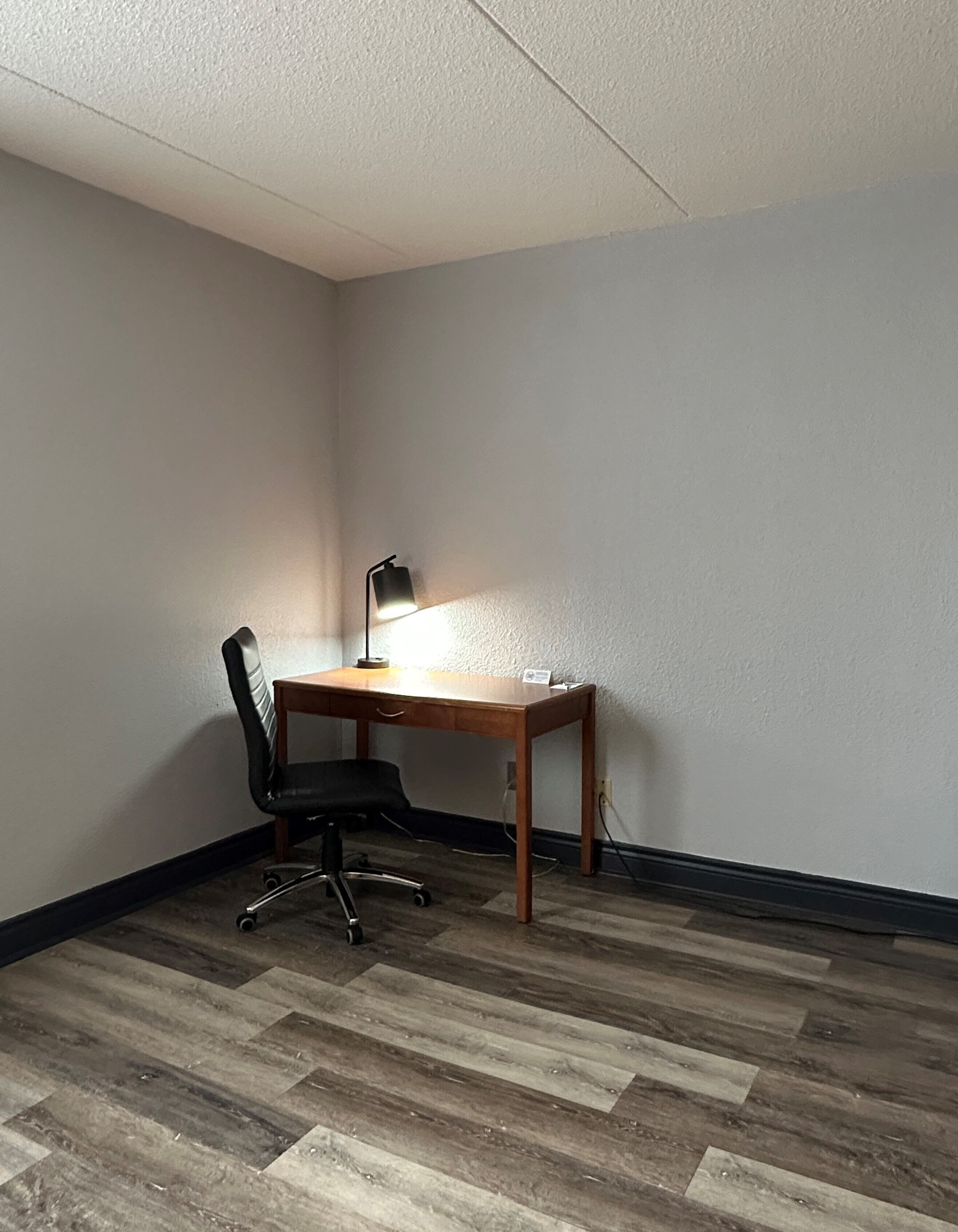 A corner room with a wooden desk, black office chair, and a desk lamp.