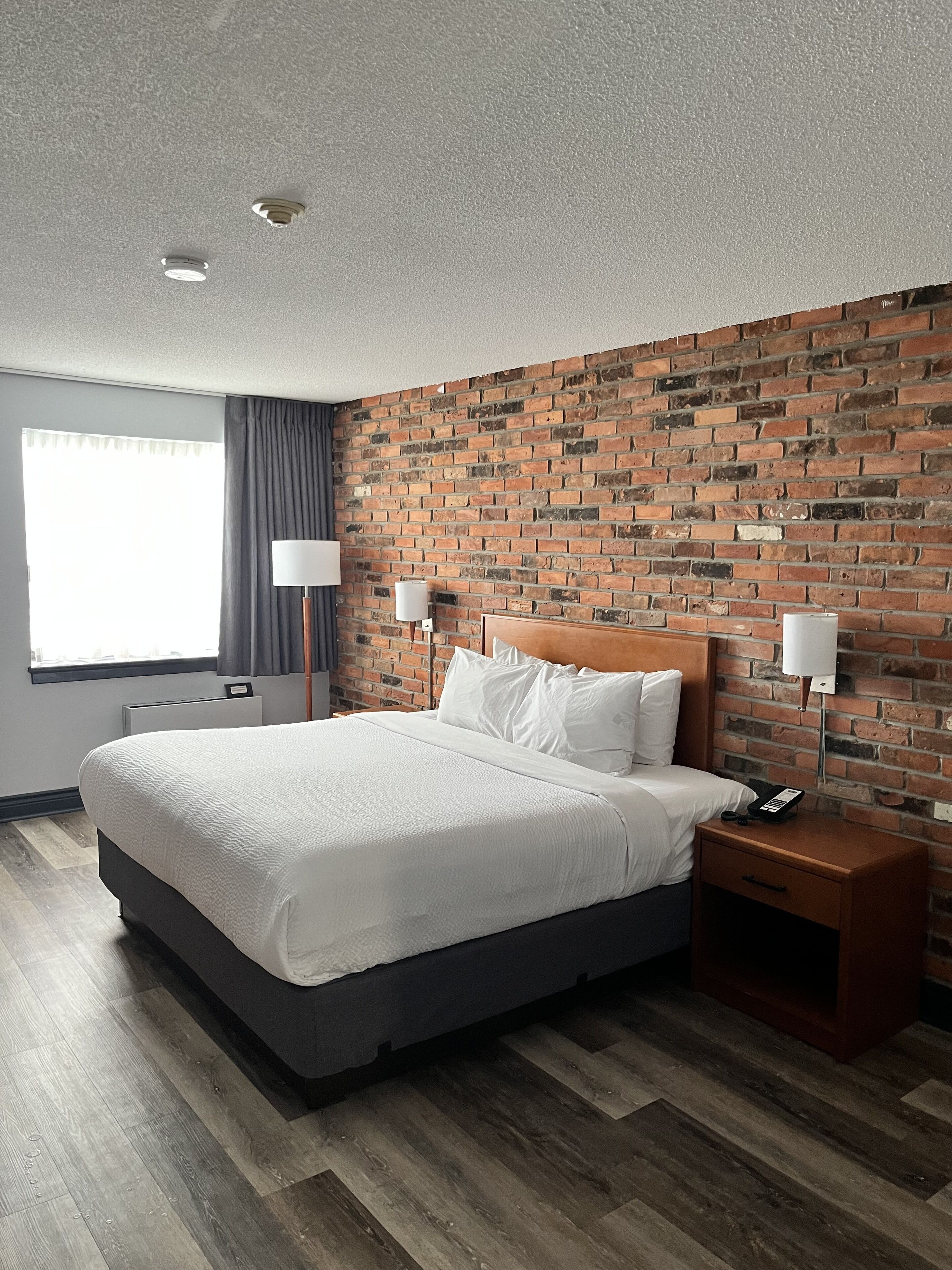 A hotel room with a brick wall, a large bed with white bedding, two bedside lamps, and a wooden nightstand.