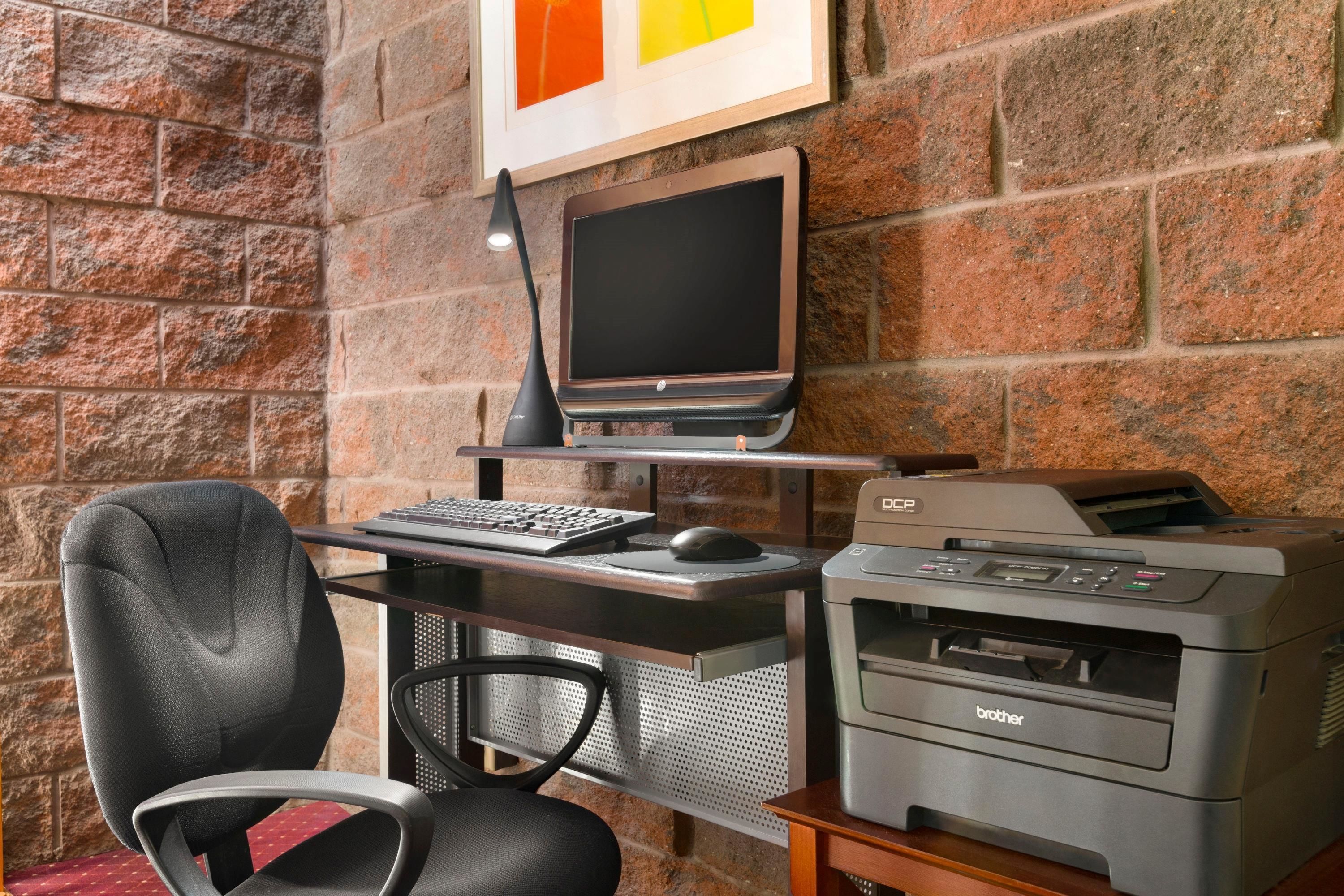 Un espacio de trabajo en casa con una computadora portátil, monitor de escritorio, impresora y una silla de oficina, todo frente a una pared de ladrillo.