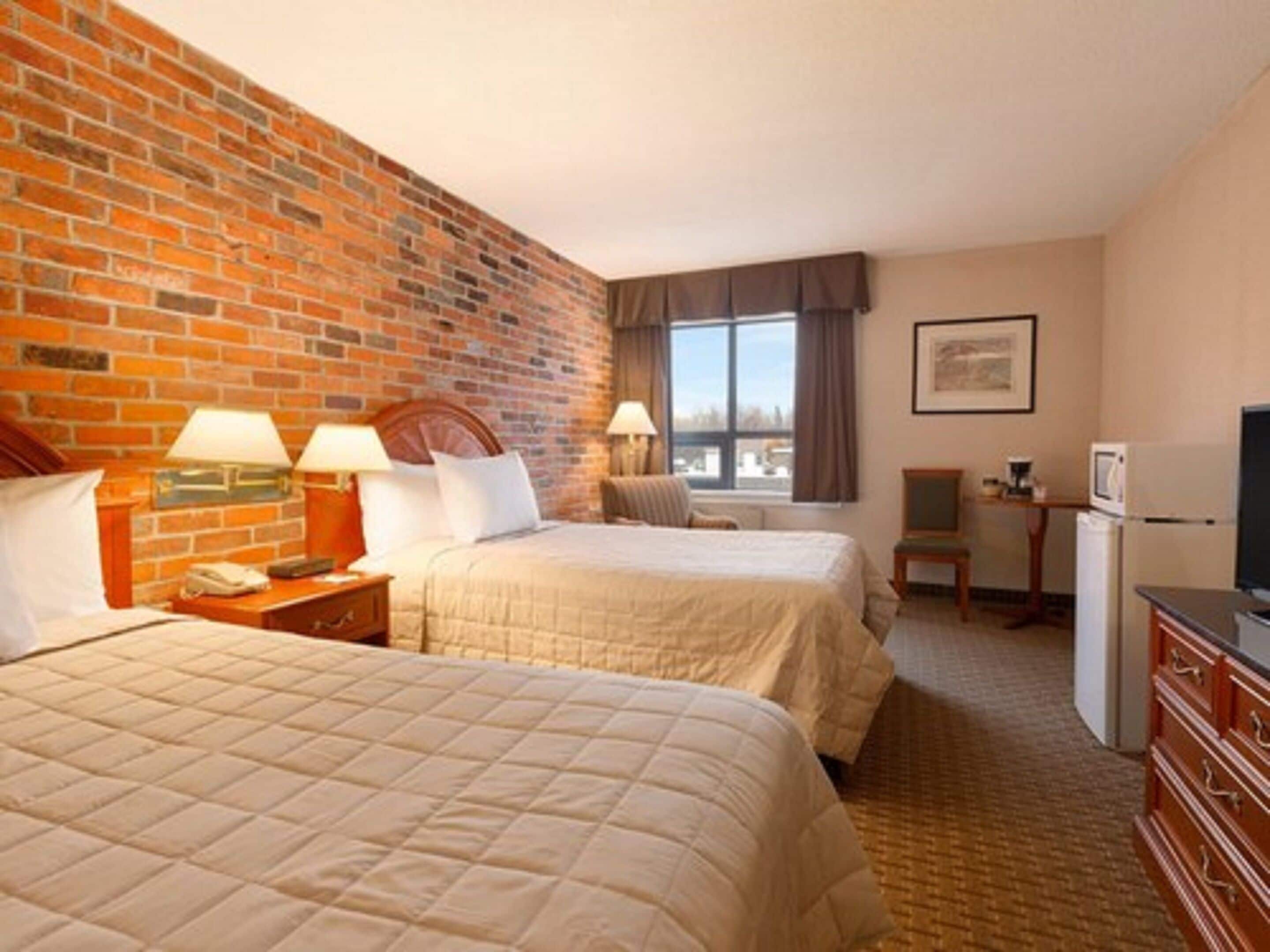 A hotel room with two beds, a brick wall, a window with curtains, a desk with a computer, and a television.