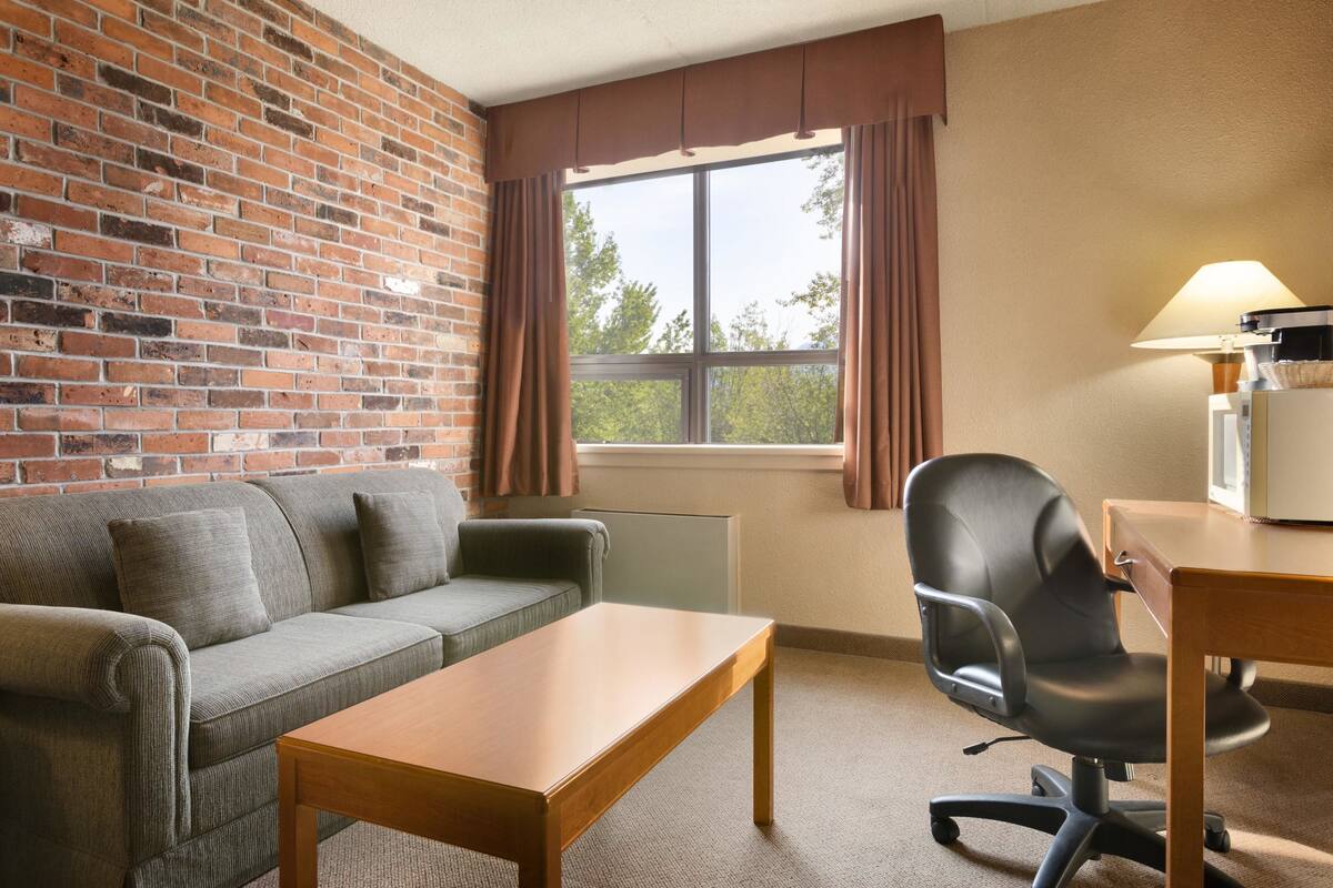 A room with a brick wall, a gray sofa, a wooden table, a black office chair, and a window with curtains.