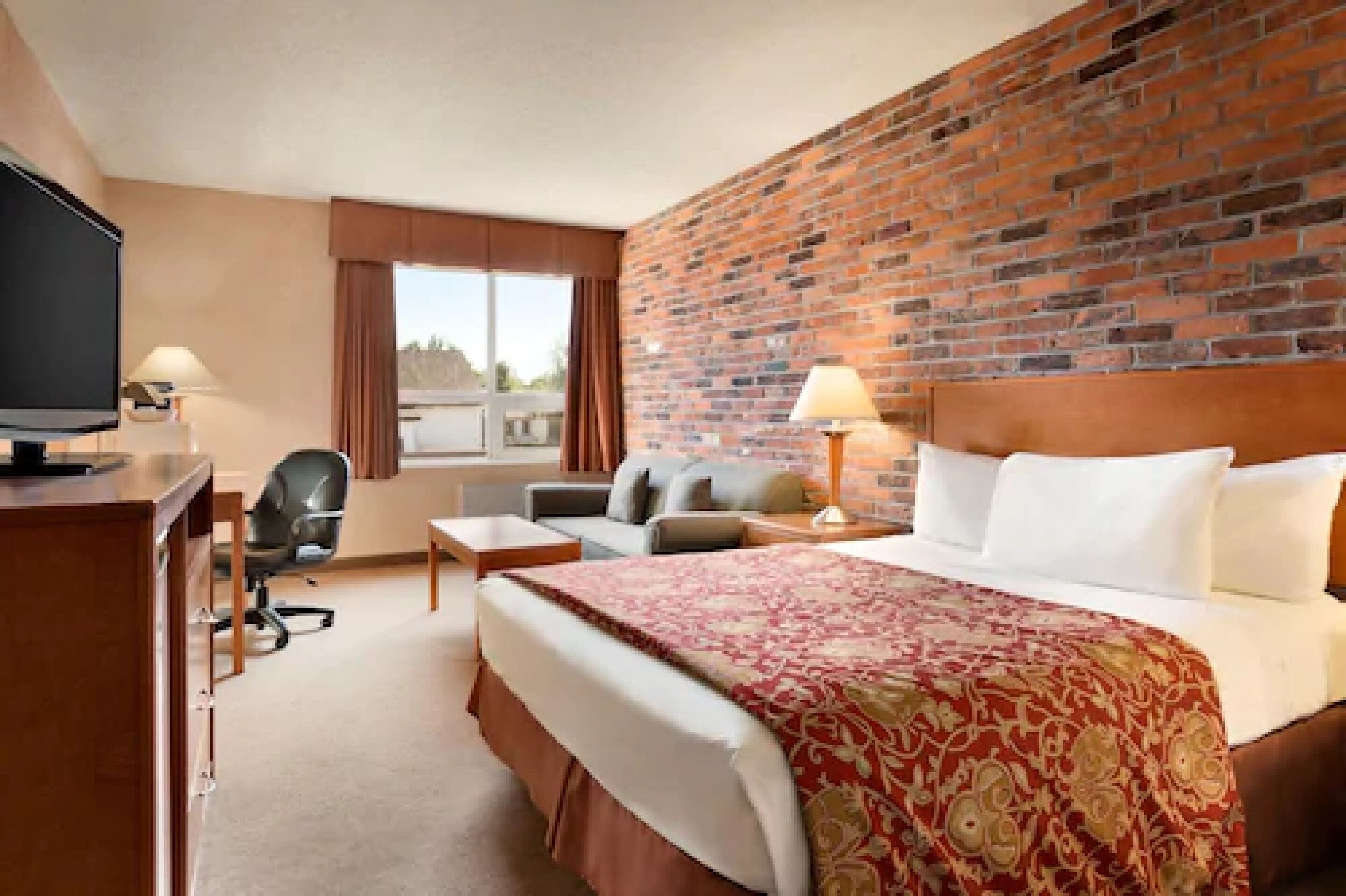 A hotel room with a brick wall, a large bed with a red patterned bedspread, a desk with a television, a chair, and a window with curtains.