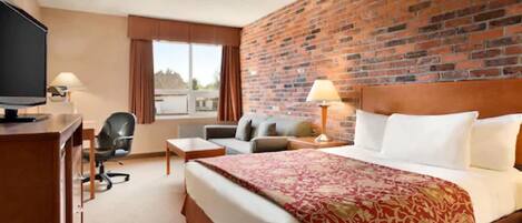 A hotel room with a brick wall, a large bed with a red patterned bedspread, a desk with a television, a chair, and a window with curtains.