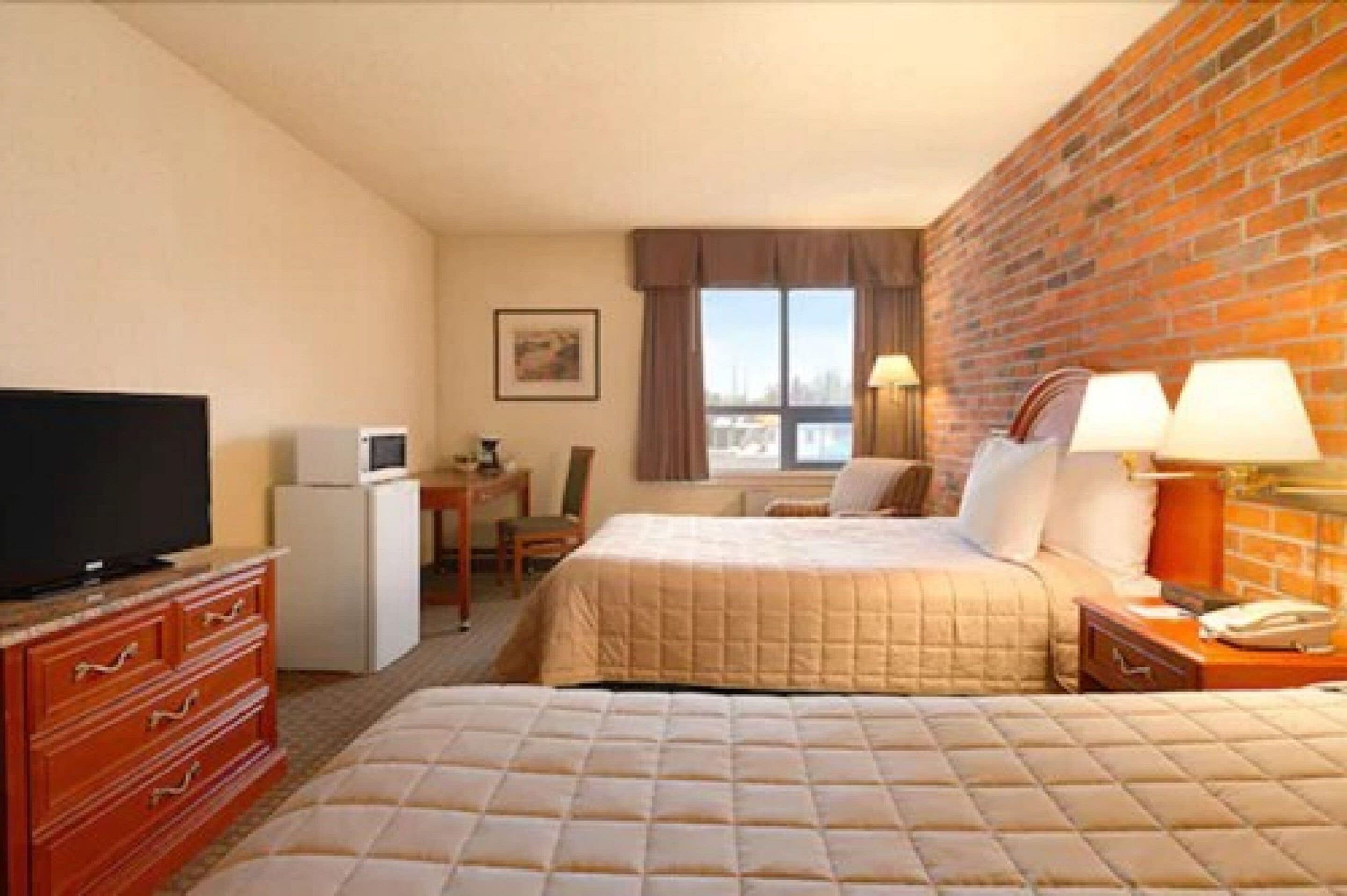 A hotel room with a brick wall, a bed, a desk with a chair, a television, and a small refrigerator.