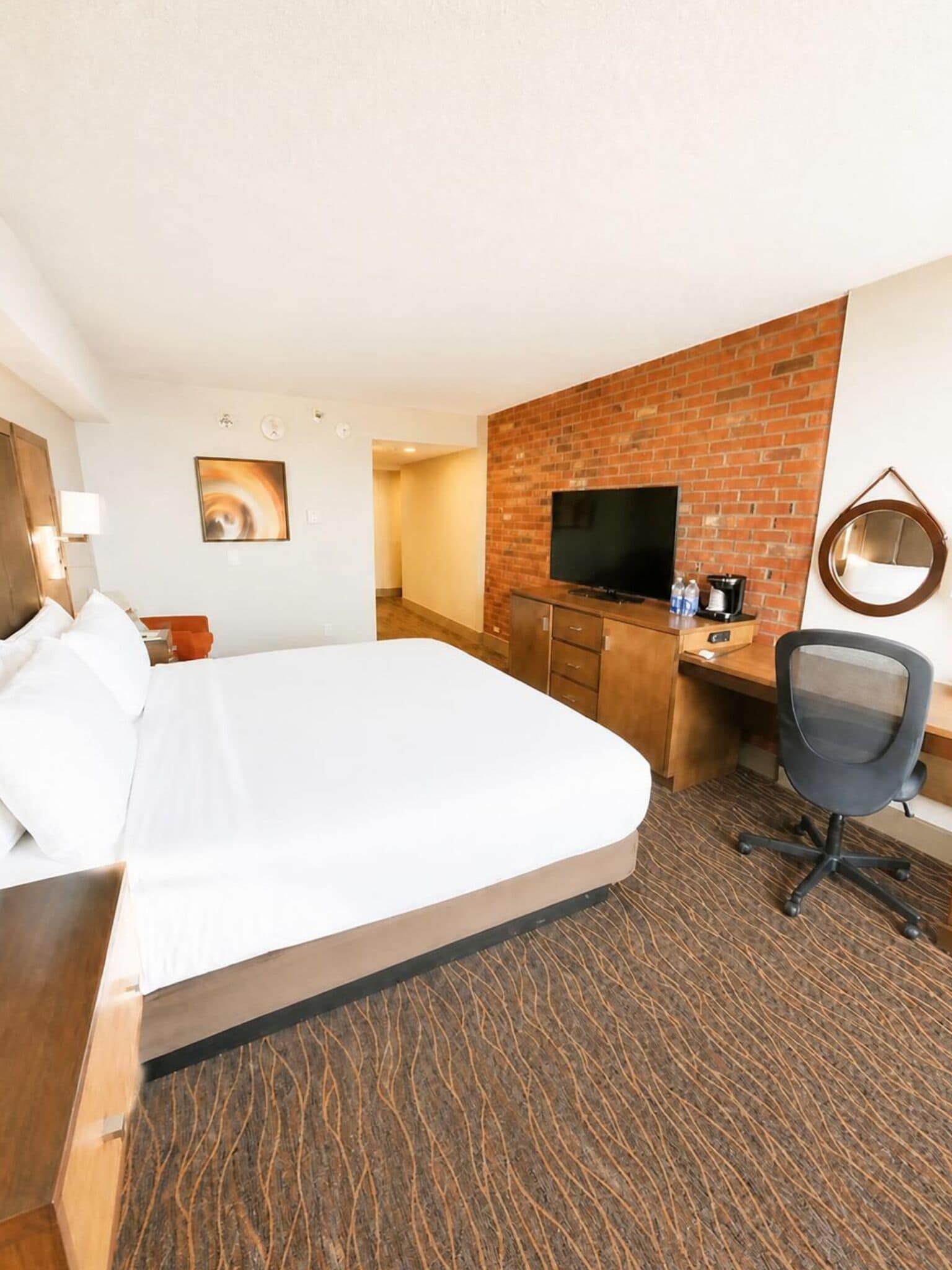 A hotel room with a bed, a desk with a TV, a chair, and a brick wall.