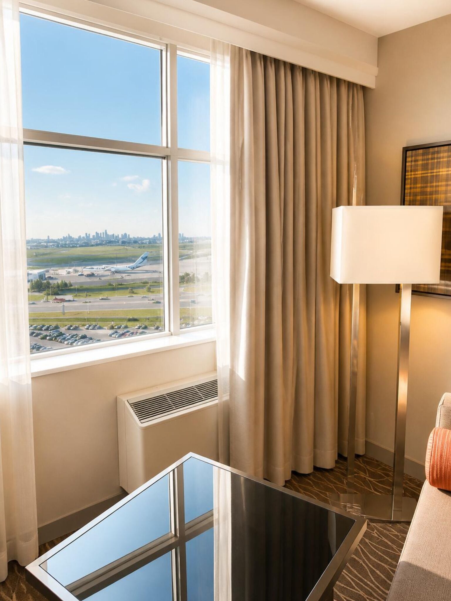A hotel room with a large window, a table, a lamp, and a view of an airport.