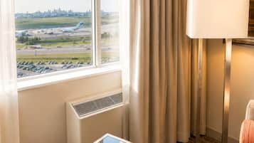 A hotel room with a large window, a table, a lamp, and a view of an airport.