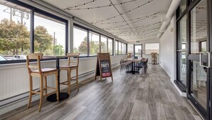 Un patio interior con sillas y mesas de madera, ventanas amplias y luces colgantes en el techo.