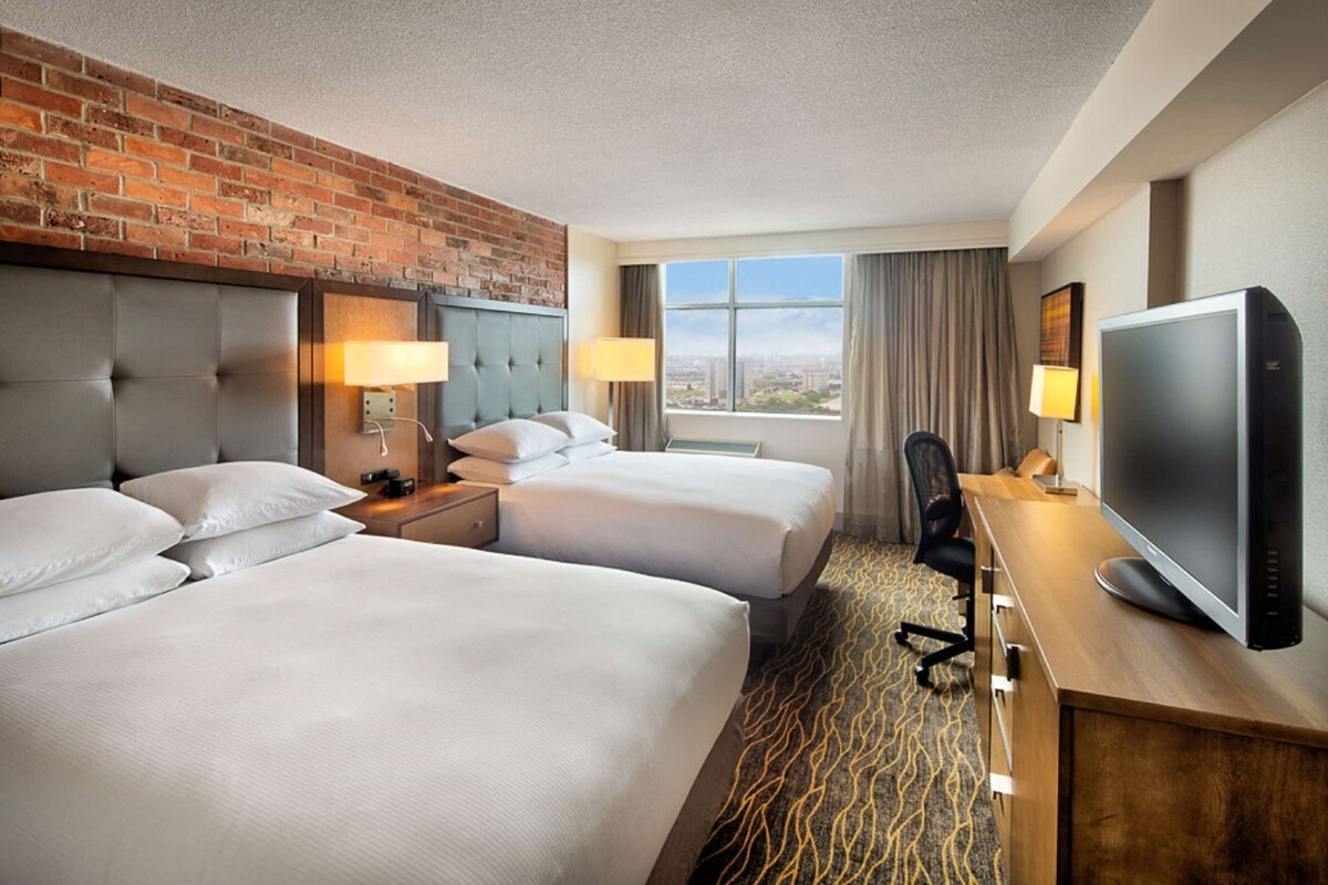 A hotel room with two beds, a TV, a desk, and a view of the city.