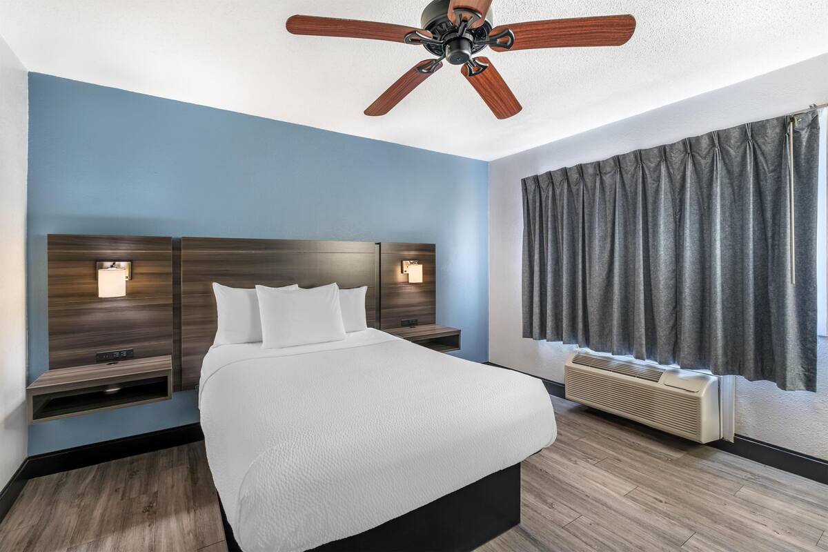 A hotel room with a bed, a ceiling fan, a wall-mounted TV, and a window with curtains.