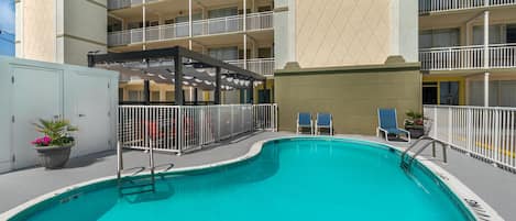 A swimming pool with a covered area, a fenced deck, and a building with multiple balconies in the background.