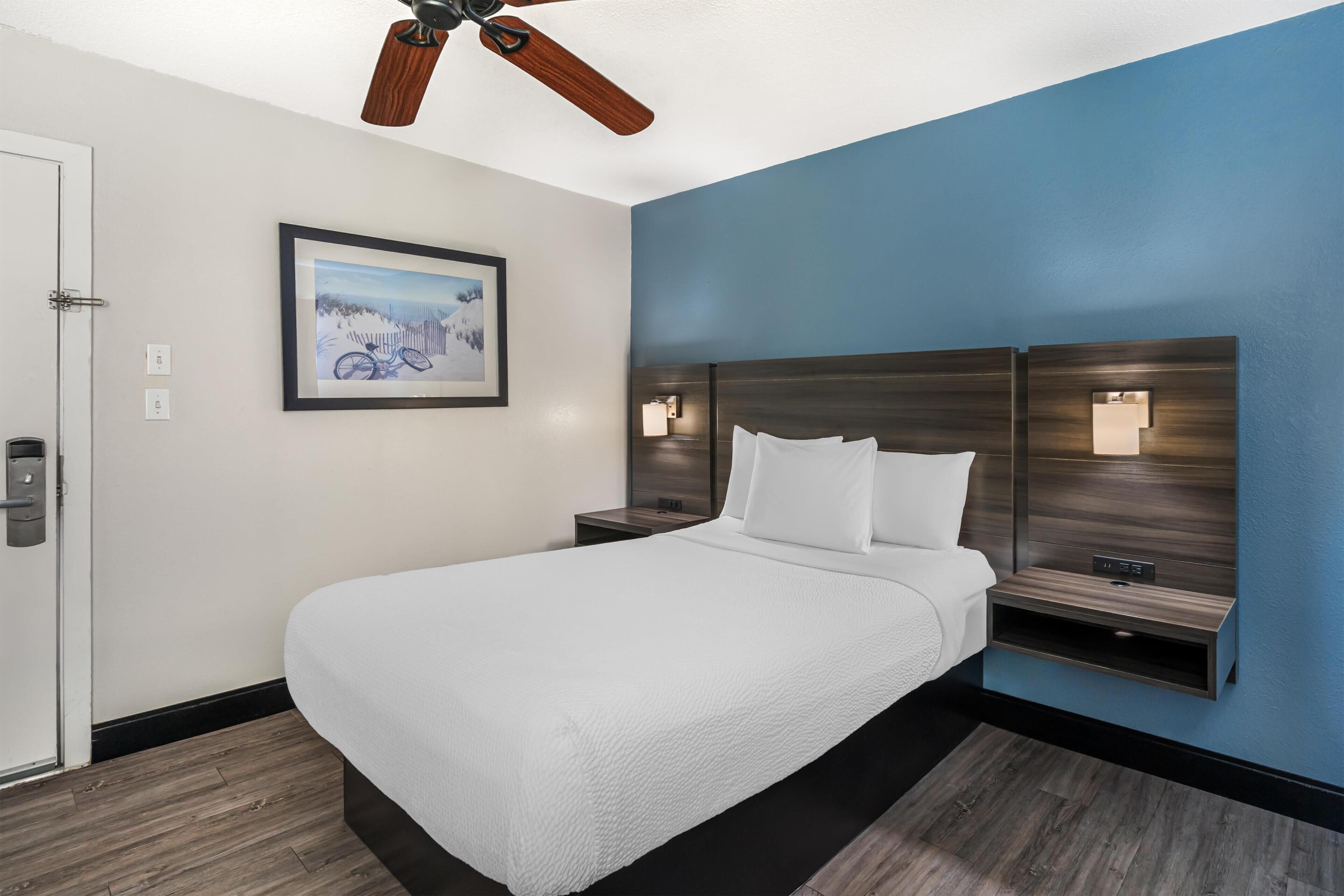 A hotel room with a bed, a ceiling fan, a framed picture on the wall, and a nightstand with a lamp.