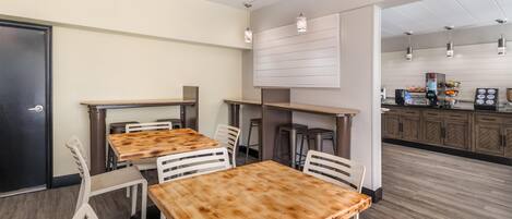Un interior moderno con mesas de madera y sillas blancas, una zona de kitchenette e iluminación colgante.